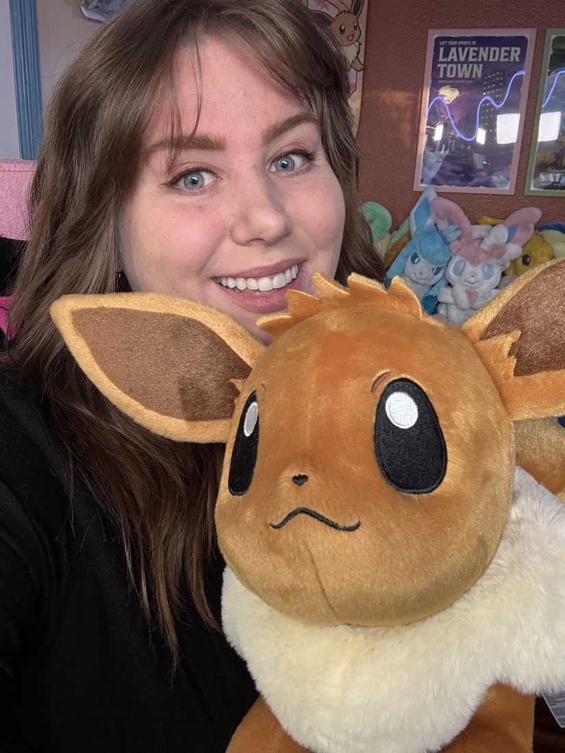 Bry smiling closely beside a large orange Eevee plush, with a shelf of Pokémon plushes and posters behind her